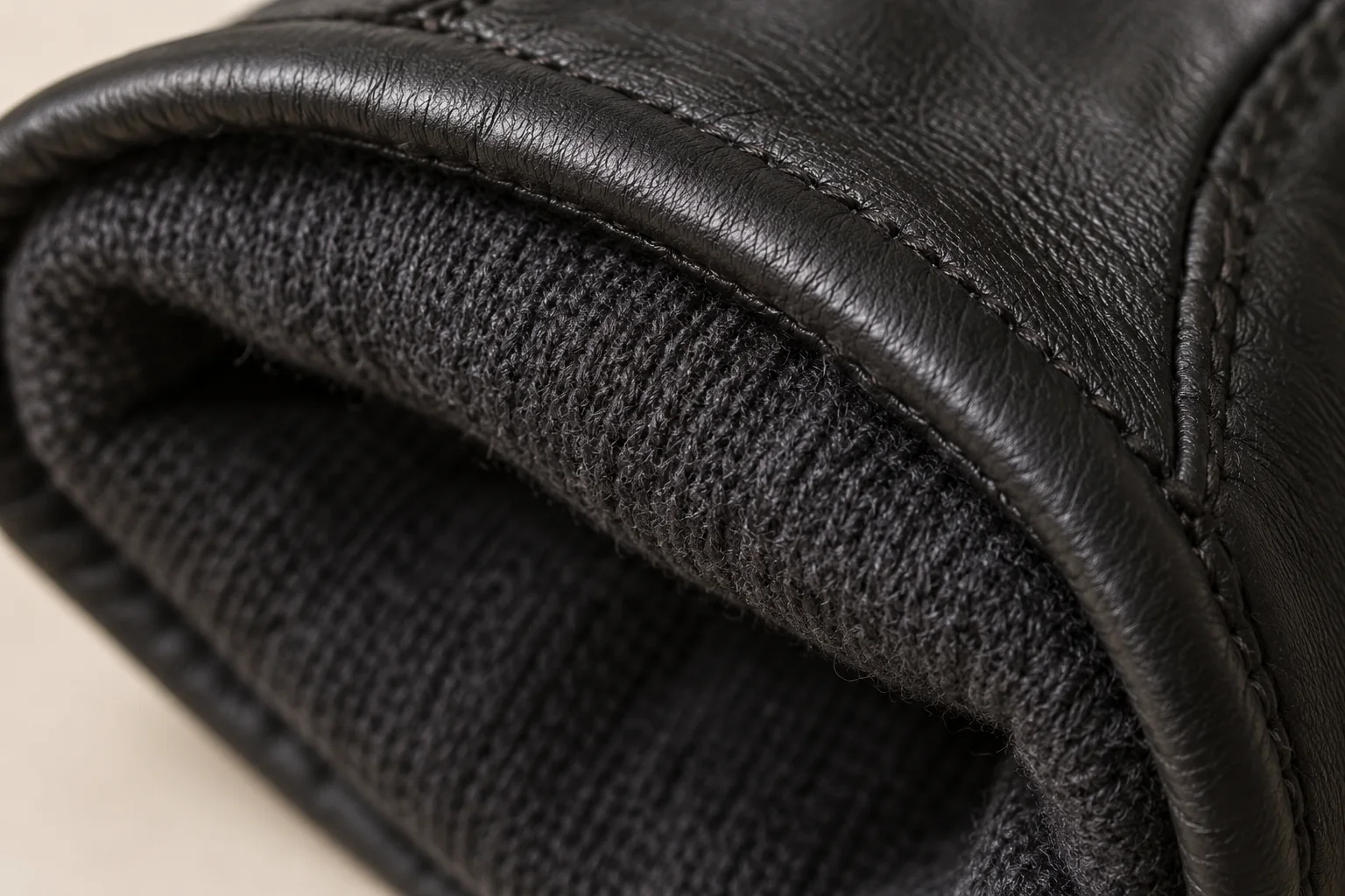 Black leather glove: tight close-up at inner cuff join showing dark charcoal fine-knit lining attached beneath a thin turned black leather rim.