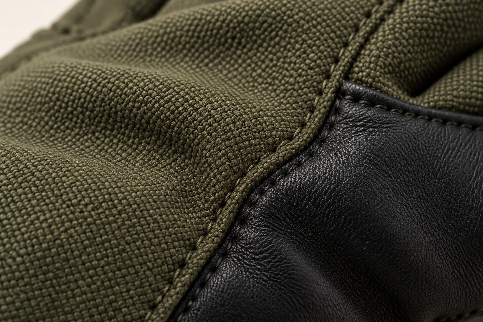 Olive woven-shell glove: macro curved hand-back side seam meeting a black smooth palm strip.