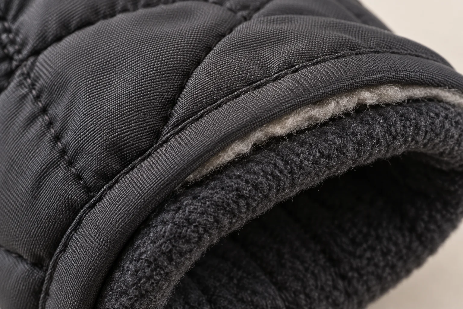 Charcoal quilted winter glove: macro cuff crossover between outer charcoal woven shell and dark fleece lining, a tiny edge of pale loft visible within turned join.